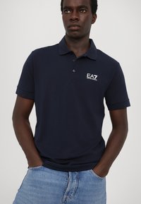 Námornícky polo tričko vyrobené z bavlny, s klasickým golierom, krátkymi rukávmi a vyšitým logom EA7 na ľavej hrudi. Nosí sa s modrými džínami.