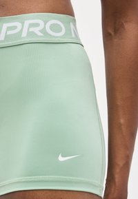 Κοντινή λήψη των ανοιχτού πράσινου σορτς Nike Pro που φοριούνται από άτομο με σκουρόχρωμο δέρμα, εμφανίζοντας το λευκό λογότυπο Nike και τη λέξη "PRO" στη μέση.