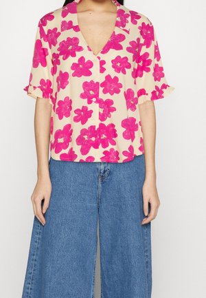 Femme portant un chemisier beige à manches courtes avec un motif floral rose vif et un jean denim bleu à jambes larges.