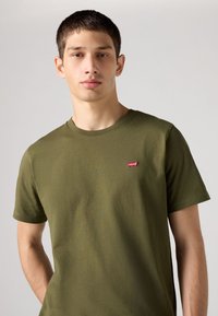 Olivno zelena bombažna majica z okroglim izrezom in kratkimi rokavi, z majhnim rdečim logotipom Levi's na levi strani prsnega koša. Gladek tekstura.