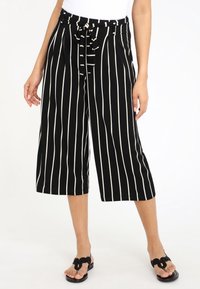 Culottes rayées noir et blanc avec une taille haute, une ceinture nouée, des jambes larges et une texture lisse ; accompagnées de sandales noires.