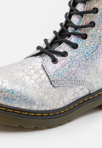 Srebrny sznurowany boot z teksturowanym, iryzującym wykończeniem. Posiada czarne sznurówki, okrągłe oczka oraz grubą podeszwę z żółtym przeszyciem.