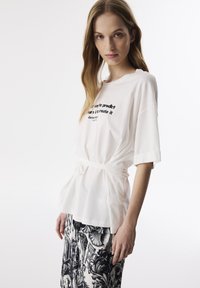 Witte t-shirt met korte mouwen en een losse pasvorm, met een zwart grafisch tekstprint. Vastgebonden rond de taille met een riem. Gecombineerd met een rok met zwart-wit patroon.