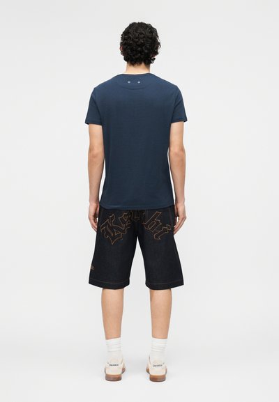 Άνδρας με σγουρά μαλλιά που φοράει μπλε σκούρο μπλουζάκι, μαύρα σορτς έως το γόνατο με πορτοκαλί γράμματα, λευκές κάλτσες και λευκά αθλητικά παπούτσια, στέκεται στραμμένος προς τα πίσω.