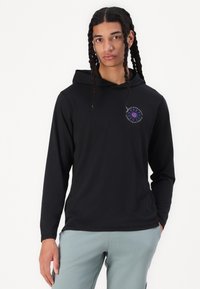 Chemise à capuche noire à manches longues avec un petit logo circulaire multicolore sur la poitrine. Texture lisse, coupe décontractée, associée à un pantalon bleu clair.