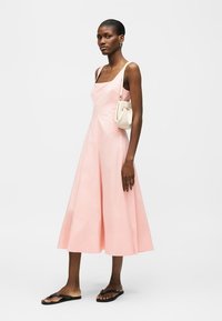 Γυναίκα που φοράει ένα ροζ μίντι φόρεμα χωρίς μανίκια, κρατώντας μια μικρή λευκή τσάντα ώμου και φορώντας μαύρα σαγιονάρια, σε όρθια στάση μπροστά σε ένα απλό φόντο.