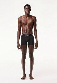 Melnas bokseršorti ar zīmola jostu, pieguloša dizaina un gluda materiāla, attēlotas uz stāvoša modeļa pret vienkāršu fonu.