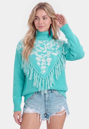 Lyshåret kvinde iført turkis højhalset sweater med hvidt mønster og frynser, kombineret med lyseblå slidte denimshorts.