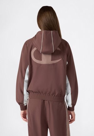 Persona que lleva una chaqueta marrón con capucha, puños elásticos y un gran logo estilizado en la espalda, combinada con pantalones marrones a juego.