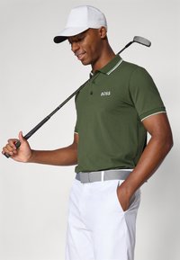 Polo verde con accenti bianchi, cintura grigia e cappellino bianco. Bastone da golf tenuto sopra la spalla, modello che indossa pantaloni bianchi. Design semplice.
