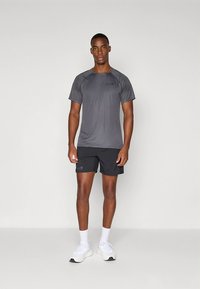 Pelēka sporta T-krekls ar īsām piedurknēm, gluda tekstūra un logotipa detaļa, kopā ar melnām šortiem un baltiem sporta apaviem.