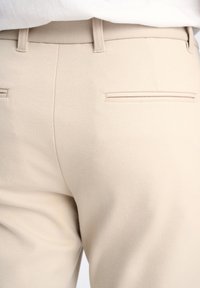 Pantaloni beige in tessuto strutturato, con due tasche posteriori e un taglio su misura. Design minimalista, senza hardware o decorazioni visibili.