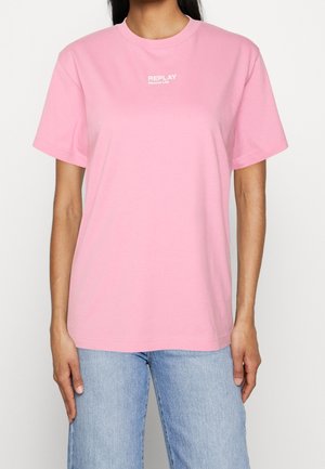 Frau trägt ein hellrosa kurzärmeliges T-Shirt mit dem Text "REPLAY Second Life", kombiniert mit hellblauen Jeans vor einem einfarbigen Hintergrund.