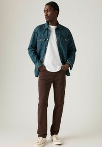 Chaqueta de mezclilla, azul profundo, con dos bolsillos en el pecho, llevada sobre una camiseta blanca. Pantalones marrones holgados y zapatillas blancas con cordones claros.