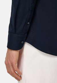 Námořnická košile s dlouhým rukávem a knoflíkovými manžetami, texturovaná látka, decentní značení na dolním lemu a bílý pozadí pro kontrast.