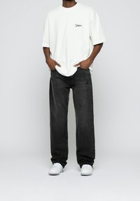 Camiseta blanca oversized con un pequeño gráfico verde en el pecho, combinada con jeans gris oscuro de corte holgado y zapatillas blancas.