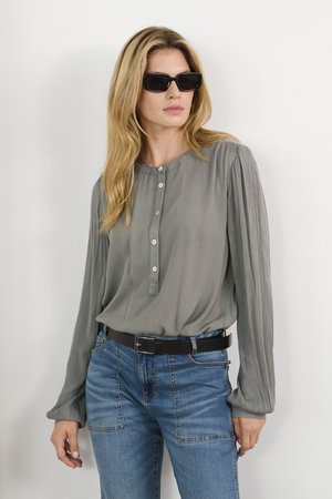 Ljusgrå blus med långa ärmar, knappraden och vertikala veck, matchad med blå jeans och ett svart bälte. Svarta solglasögon kompletterar stilen.