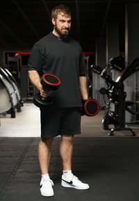 Zwarte T-shirt met korte mouwen en shorts, houdt dumbbells vast met rode accenten, witte sneakers. Sportschoolomgeving met fitnessapparatuur op de achtergrond.