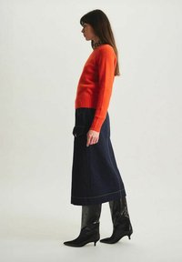 Pull en laine orange vif, jupe longue en denim foncé avec une poche, et bottes noires vernies jusqu'au genou avec un bout pointu.