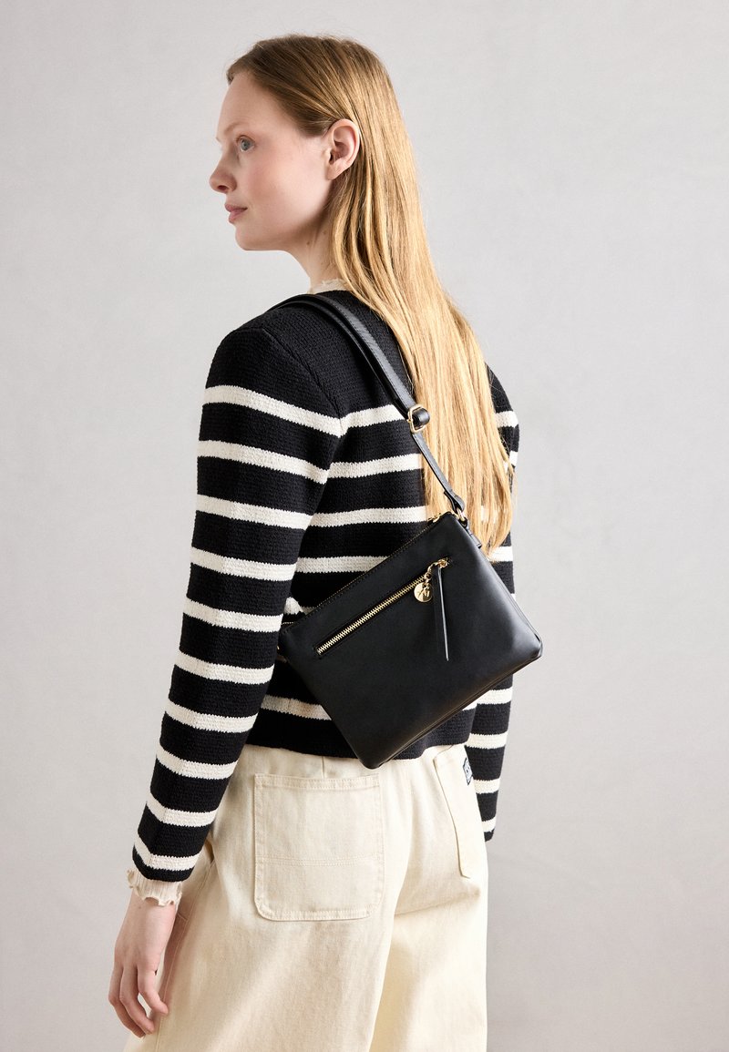 Bolso cruzado de cuero negro con un bolsillo frontal con cremallera y detalles de hardware en dorado. La modelo lleva puesto un suéter de rayas en blanco y negro.