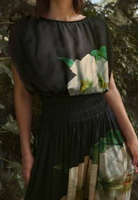 Femme portant une robe noire avec de grandes impressions florales blanches, taille cintrée et jupe froncée, se tenant à l'extérieur près de feuillages verts.