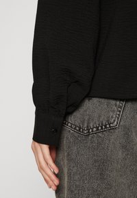Black textured long-sleeve shirt with a gathered cuff detail, fastened by a black button, paired with gray denim jeans featuring contrasting stitching.