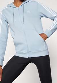 Sudadera con cremallera de color azul claro, con rayas blancas a lo largo de las mangas, bolsillos frontales y capucha ajustable con cordón. Presenta un pequeño logo en el pecho.