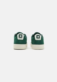 Adidași din suede verde cu tălpi din cauciuc alb. Au un patch circular cu logo pe călcâi, care include o stea și textul "CONVERSE".