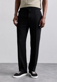 Czarne, dopasowane spodnie z elastycznym paskiem. Gładki materiał o prostym kroju. Połączone z czarnymi trampkami z białymi podeszwami i sznurowadłami.