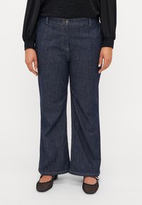 Wide-leg dark denim jeans with a zip and button closure, featuring dual front pockets and a relaxed fit. Complemented by flat, mesh-textured shoes.