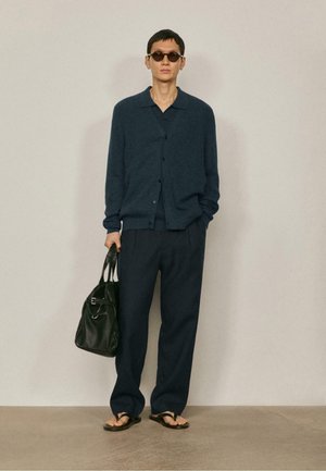 Mann trägt dunkelblauen Cardigan, passende Hose, Sonnenbrille, Sandalen, hält schwarze Ledertasche, steht vor einer einfarbigen hellgrauen Wand.