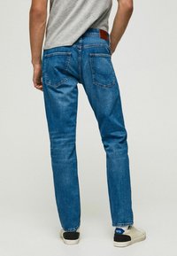 Blå jeans i denim med rak benpassform, utrustade med två bakfickor och en brun läderpatch vid midjan, kombinerade med ljusa sneakers.