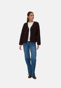 Veste en polaire marron texturé avec un col en V, fermeture à boutons et poches latérales, portée sur une chemise blanche et un jean évasé bleu.