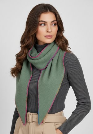 Woman with wavy brown hair wearing a gray ribbed turtleneck, beige belted pants, and a green triangular scarf with colorful stitched edges.