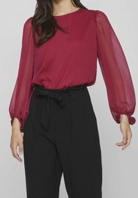 Donna che indossa una blusa rossa trasparente a maniche lunghe infilata in pantaloni neri a vita alta con una cintura annodata in vita.