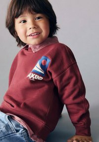 Felpa bordeaux con un logo blu e rosso che dice "WEEKEND", abbinata a una camicia color rosa chiaro con colletto e jeans in denim. Capelli castani, sorridente.