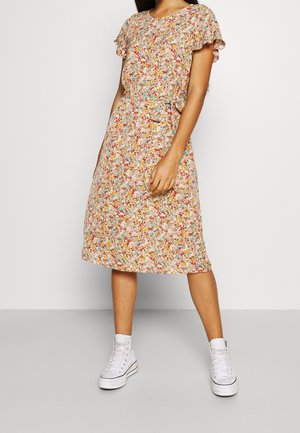 Femme portant une robe fleurie longueur genou à manches courtes associée à des baskets montantes blanches sur un fond uni.