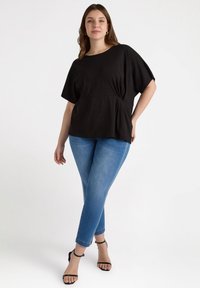 Top negro de mangas cortas con diseño de cintura ajustada, combinado con jeans ajustados azules y sandalias negras de tiras sobre un fondo claro.