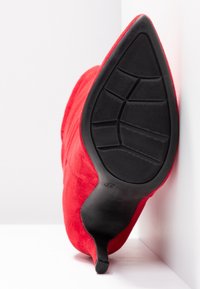 Escarpin en daim rouge à bout pointu vu de la semelle, montrant une semelle extérieure en caoutchouc noir texturé et un talon fin.