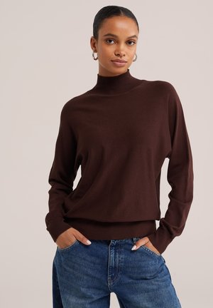 Frau mit Creolen, die einen dunkelbraunen Rollkragenpullover und blaue Jeans trägt, mit den Händen in den Taschen, neutraler Hintergrund.