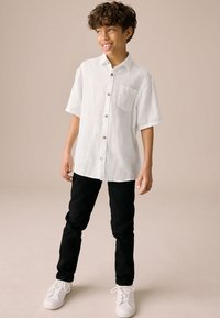 Weißes Leinen-Kurzarmhemd mit einer Tasche und Holzknöpfen, kombiniert mit schwarzen Hosen und weißen Sneakers. Einfaches, lässiges Outfit.