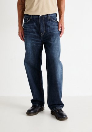 Dunkelblaue Denim-Jeans mit geradem Schnitt, dezentem Verblassen und kontrastierenden Nähten. Kombiniert mit schwarzen Lederschuhen.