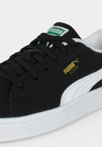 Κοντινό πλάνο μαύρου σουέτ αθλητικού Puma με λευκή πλαϊνή ρίγα, λευκή ανάγλυφη σόλα και πράσινο λογότυπο Puma στη γλώσσα.