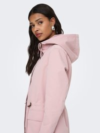 Femme aux longs cheveux foncés portant des boucles d'oreilles argentées en forme de cercle et une veste à capuche rose clair, vue de profil sur un fond blanc.