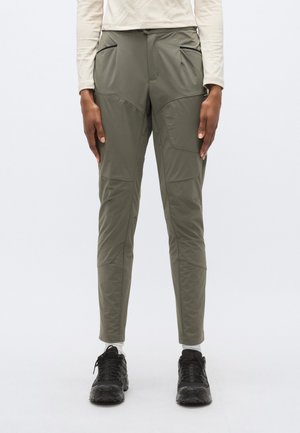 Olívovo zelené nohavice s mäkkou škrupinou, úzkym strihom, viacerými vreckami a zosilneným dizajnom na kolenách. Kombinované s čiernymi teniskami.