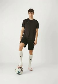 Noor poiss mustas Puma sportlikus riietuses seisab ühe jalaga jalgpallil, kandes valgeid sokke ja jalatseid tasas taustal.