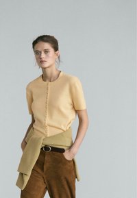 Short-sleeve, buttoned, light yellow knit top paired with a beige, textured knit wrap and brown suede pants, detailed with a brown belt.
