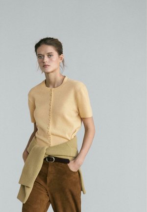 Maglia a maniche corte giallo chiaro con bottoni, abbinata a un avvolgente beige lavorato a maglia dalla trama, e pantaloni in suede marrone, completati da una cintura marrone.