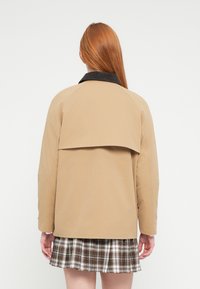 Ταμπά σακάκι με μαύρο γιακά, με σχεδιασμό σε επίπεδα στην πλάτη. Φορεμένο πάνω από μια καρό φούστα, παρουσιάζει απαλή υφή και δομημένη εφαρμογή.