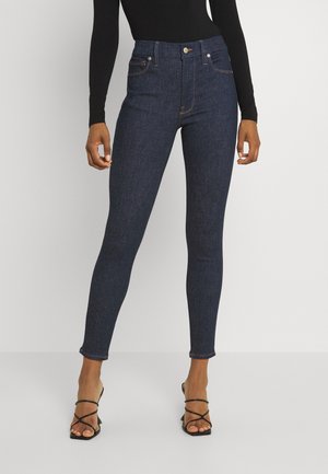 Nainen pukeutuneena tummansinisiin skinny-farkkuihin, mustaan pitkähihaiseen paitaan ja mustiin remmikkäisiin korkokenkiin, seisomassa valkoista taustaa vasten.